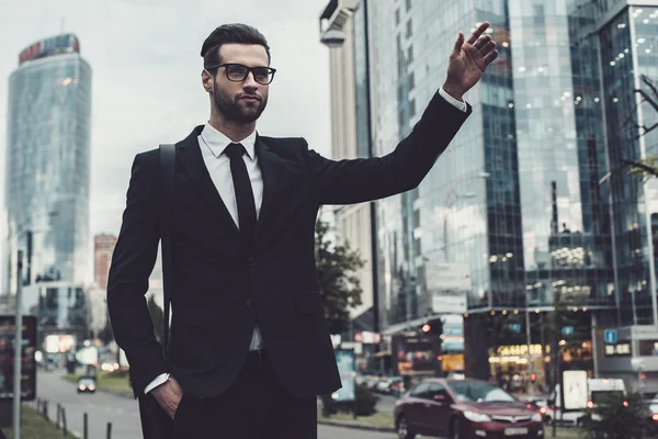 ¡Taxi! Joven empresario confiado en traje completo cogiendo un taxi mientras levanta el brazo y se para al aire libre con paisaje urbano en el fondo — Foto de Stock