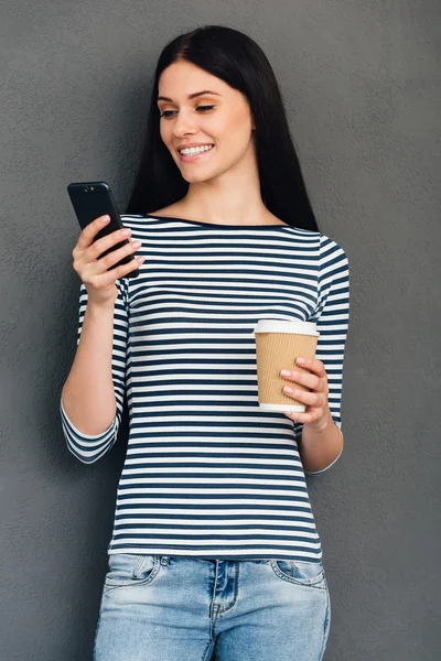Frau hält Kaffeetasse in der Hand und blickt auf ihr Smartphone — Stockfoto