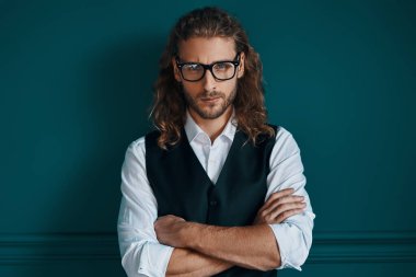 Zarif giyinmiş yakışıklı genç adam kameraya bakıyor ve yeşil arka planda dururken kollarını çapraz tutuyor.