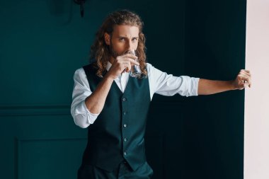 Zarif giyinmiş yakışıklı genç adam yeşil arka planda dikilirken kameraya bakıyor ve su içiyor.