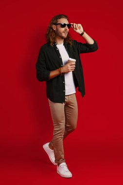 Kırmızı arka planda dikilirken gülümseyerek göz giysisini ayarlayan sıradan giyinmiş, tam boy modaya uygun bir genç.