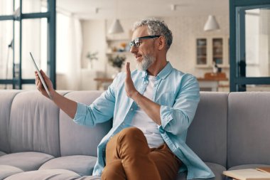 Modern senior man using digital tablet and waving hand while sitting on the sofa at home