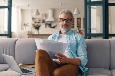 Busy senior man checking the papers and using laptop while sitting on the sofa at home