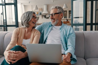 Happy senior couple in casual clothing smiling and using laptop while bonding together at home