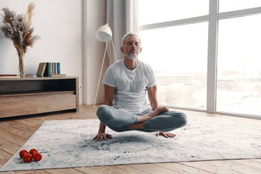 Active senior man in sport clothing meditating while standing on hands in the yoga position at home