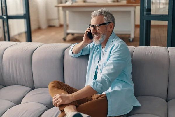 Happy senior man in casual clothing and eyeglasses talking on the smartphone while sitting on the sofa at home
