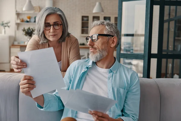 Busy senior couple in casual clothing doing paperwork while bonding together at home
