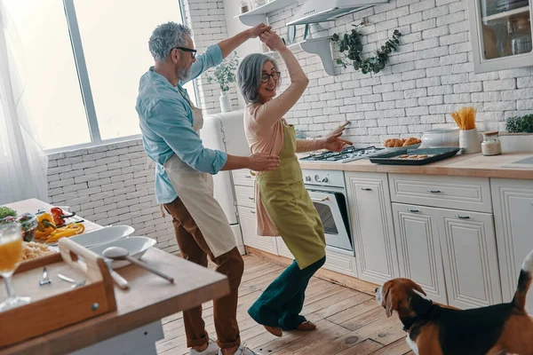 Full length of senior couple in aprons dancing and smiling while preparing healthy dinner at home