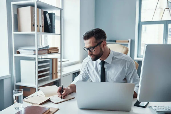 Geconcentreerde jongeman in hemd en das iets opschrijven terwijl hij op kantoor zit — Stockfoto