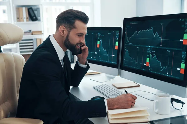 Geconcentreerde jongeman in hemd en das iets opschrijven en praten aan de telefoon terwijl hij op kantoor werkt — Stockfoto