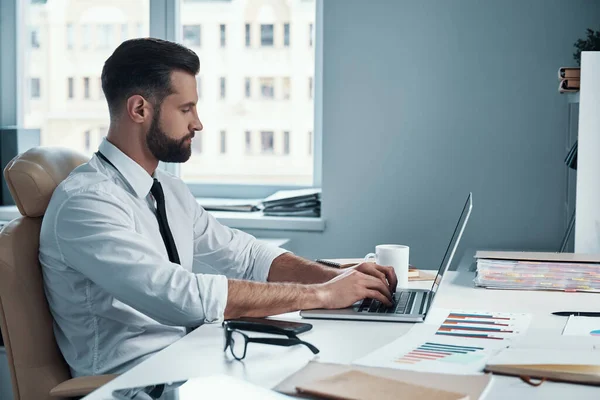 Joven concentrado en camisa y corbata trabajando en la computadora mientras está sentado en la oficina — Foto de Stock