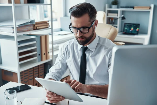 Drukke jongeman in hemd en stropdas werkend met draadloze technologieën terwijl hij op kantoor zit — Stockfoto