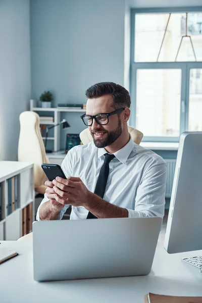 Bello giovane in abbigliamento formale con smart phone e sorridente mentre seduto in ufficio — Foto Stock