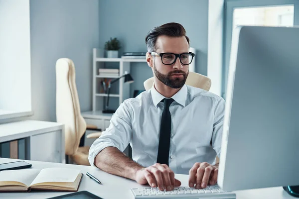 Drukke jongeman in shirt en stropdas werken op de computer tijdens het zitten in het kantoor — Stockfoto