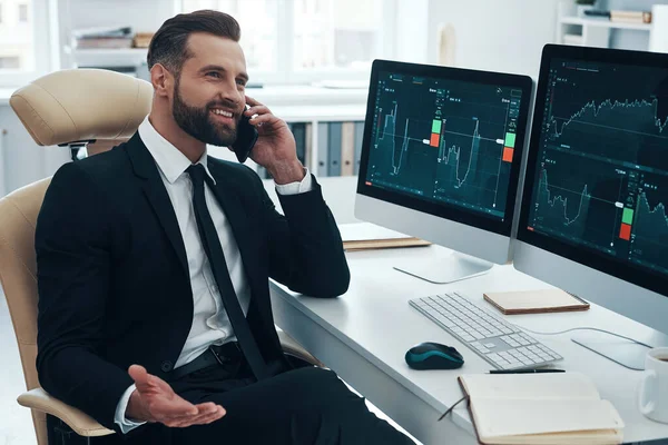 Uśmiechnięty młodzieniec w koszuli i krawacie analizujący dane na giełdzie i rozmawiający przez telefon podczas pracy w biurze — Zdjęcie stockowe