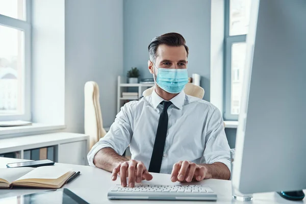 Уверенный молодой бизнесмен в защитной маске работает за компьютером, сидя в офисе — стоковое фото