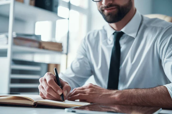 Close up of young man in formalwear writing something down while sitting in the office — Stock Photo, Image