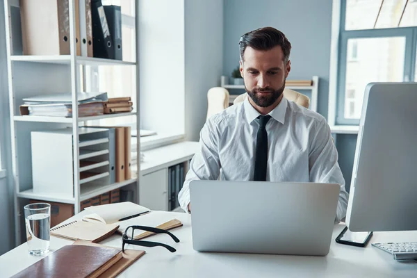 Geconcentreerde jonge man in shirt en stropdas werken met behulp van de computer tijdens het zitten in het kantoor — Stockfoto