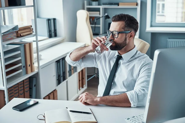Joven guapo con camisa y corbata bebiendo agua mientras está sentado en la oficina —  Fotos de Stock