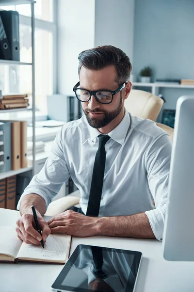 Elegant ung man i formalwear skriva något i anteckningsblock när du sitter på kontoret — Stockfoto