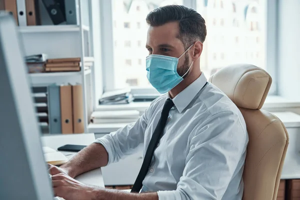 Joven empresario confiado en la máscara facial protectora que trabaja en la computadora mientras está sentado en la oficina —  Fotos de Stock