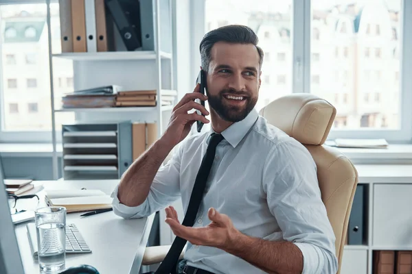 Resmi giyinmiş yakışıklı genç adam telefonda konuşuyor ve ofiste otururken gülümsüyor. — Stok fotoğraf