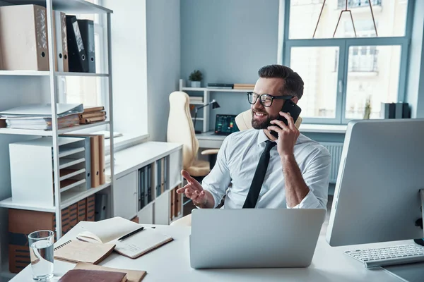 Guapo joven en ropa formal hablando por teléfono y sonriendo mientras está sentado en la oficina — Foto de Stock