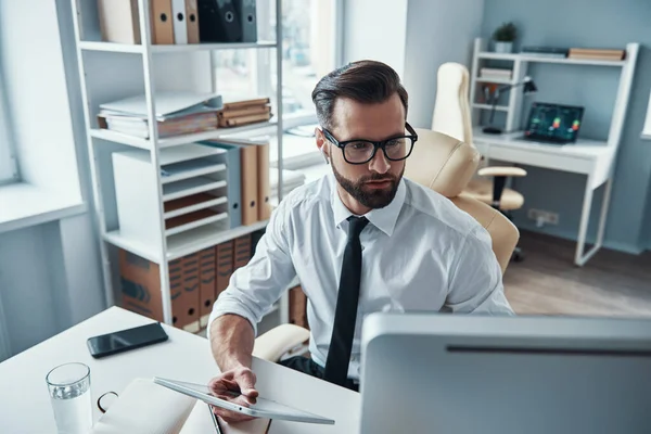 Geconcentreerde jongeman in hemd en das werkend met behulp van draadloze technologieën terwijl hij op kantoor zit — Stockfoto