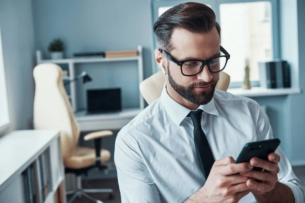 Hezký mladý muž ve formalwear pomocí chytrého telefonu a nošení sluchátek, zatímco sedí v kanceláři — Stock fotografie