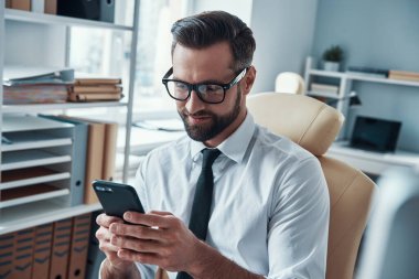 Formalite giymiş yakışıklı genç adam akıllı telefon kullanıyor ve ofiste otururken kulaklık takıyor.