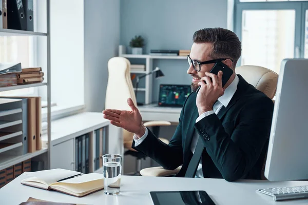 Jovem elegante de camisa e gravata falando no telefone inteligente e sorrindo enquanto está sentado no escritório — Fotografia de Stock