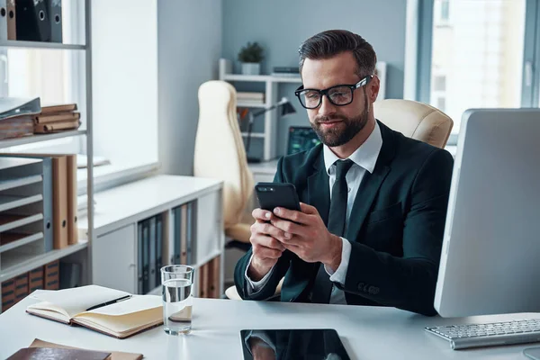 Good looking young man in shirt and tie using smart phone and smiling while sitting in the office — Stock Photo, Image