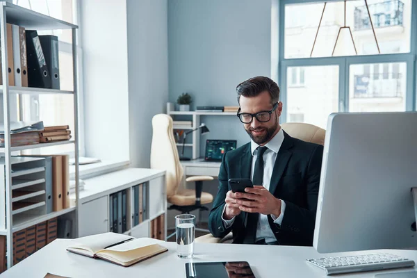 Hombre joven guapo en camisa y corbata usando teléfono inteligente y sonriendo mientras está sentado en la oficina — Foto de Stock