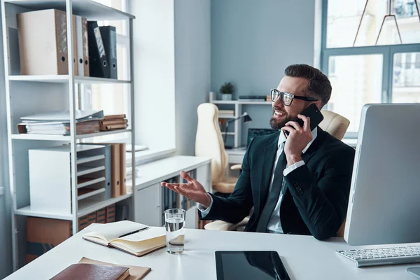 Hombre joven guapo en camisa y corbata hablando en el teléfono inteligente y sonriendo mientras está sentado en la oficina — Foto de Stock