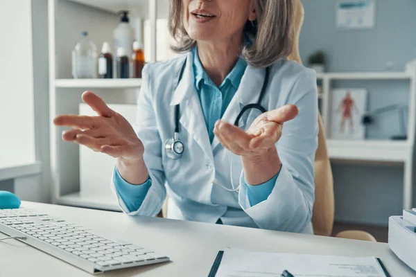 Close up de médico feminino maduro em jaleco branco dizendo algo e gesto enquanto sentado em seu escritório — Fotografia de Stock