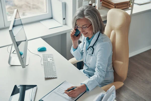 Visão superior do belo médico feminino maduro em jaleco branco falando no telefone e sorrindo enquanto está sentado em seu escritório — Fotografia de Stock