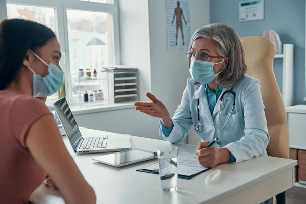 Mature female doctor in white lab coat making a prescription to young woman while sitting in her office — Stock Photo, Image