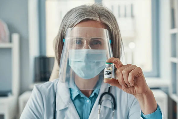 Doctora madura en bata de laboratorio blanca y ropa de trabajo protectora que sostiene una vacuna Covid-19 mientras está sentada en el consultorio médico — Foto de Stock