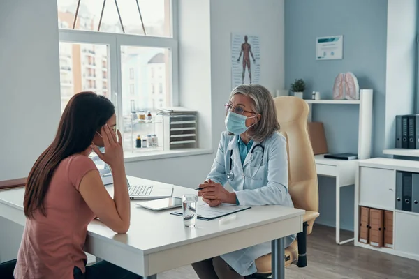 Mature female doctor in white lab coat making a prescription to young woman while sitting in her office — Stock Photo, Image