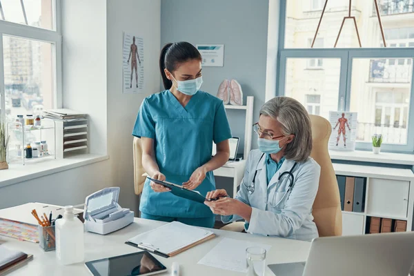 Красивая молодая медсестра в защитной маске разговаривает со зрелым врачом во время работы в больнице — стоковое фото