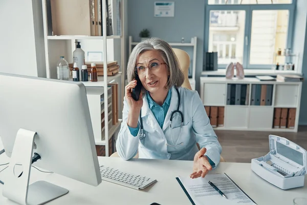Médica madura confiante em jaleco branco falando ao telefone e sorrindo enquanto estava sentada em seu escritório — Fotografia de Stock