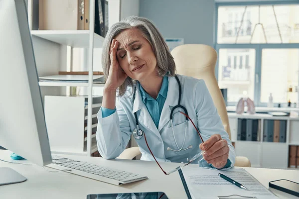 Cansado maduro feminino médico em jaleco branco mantendo os olhos fechados enquanto sentado em seu escritório — Fotografia de Stock