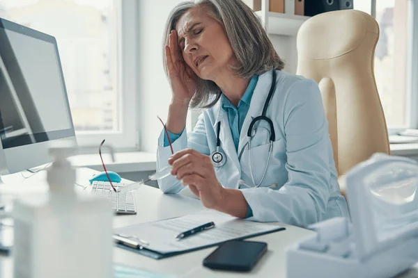 Overworked mature female doctor in white lab coat suffering from headache while sitting in her office — Stock Photo, Image