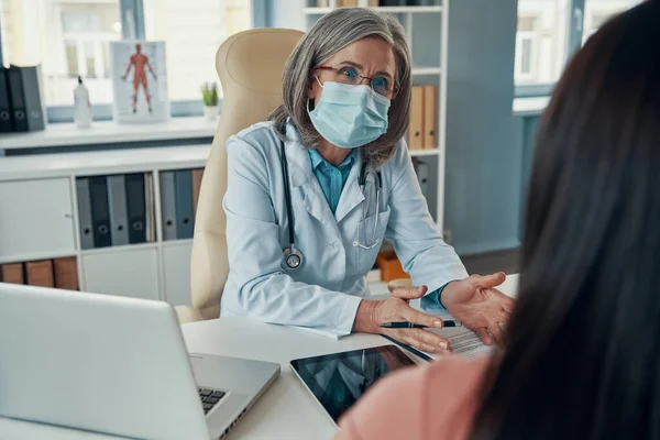 Confident female doctor in white lab coat giving a healthcare consultation to young woman while sitting in her office — Stock Photo, Image