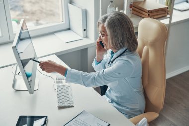 Beyaz laboratuvar önlüklü, güzel, olgun bir kadın doktorun ofisinde otururken telefonda konuşmasının ve gülümsemesinin en güzel görüntüsü.