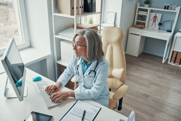 Visão superior do médico feminino maduro em jaleco branco trabalhando no computador enquanto sentado em seu escritório — Fotografia de Stock