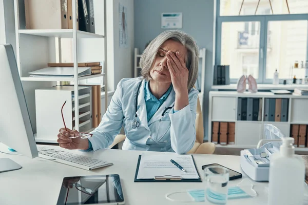 Zmęczona dojrzała lekarka w białym fartuchu, zakrywająca twarz dłonią podczas siedzenia w biurze — Zdjęcie stockowe
