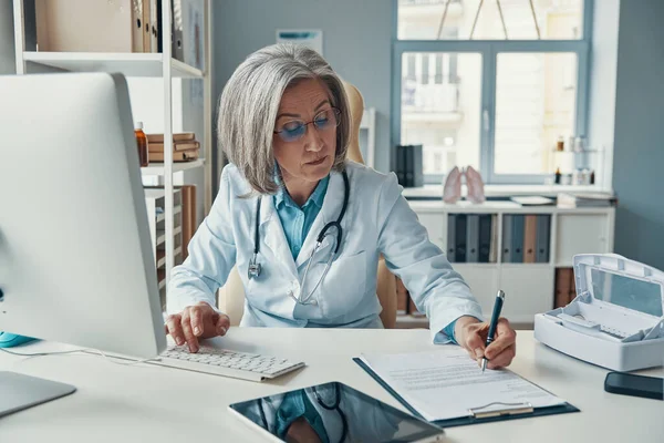 Poważna, dojrzała lekarka w białym fartuchu, pisząca coś podczas siedzenia w biurze. — Zdjęcie stockowe