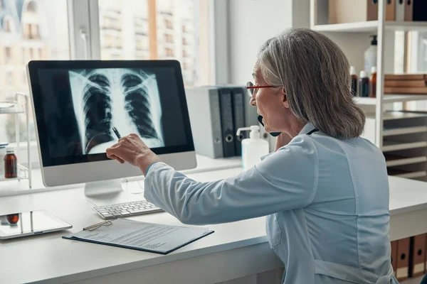 Dojrzała lekarz w białym fartuchu rozmawia przez telefon i bada płuca za pomocą komputera podczas siedzenia w biurze — Zdjęcie stockowe