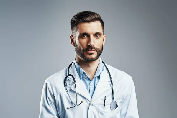 Handsome young man in white lab coat looking at camera while standing against grey background — Stock Photo, Image